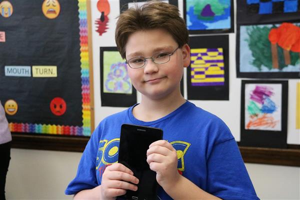 Male student holds electronic device in front of a bulletin board. Male student holds electronic device in front of a bulletin board.
